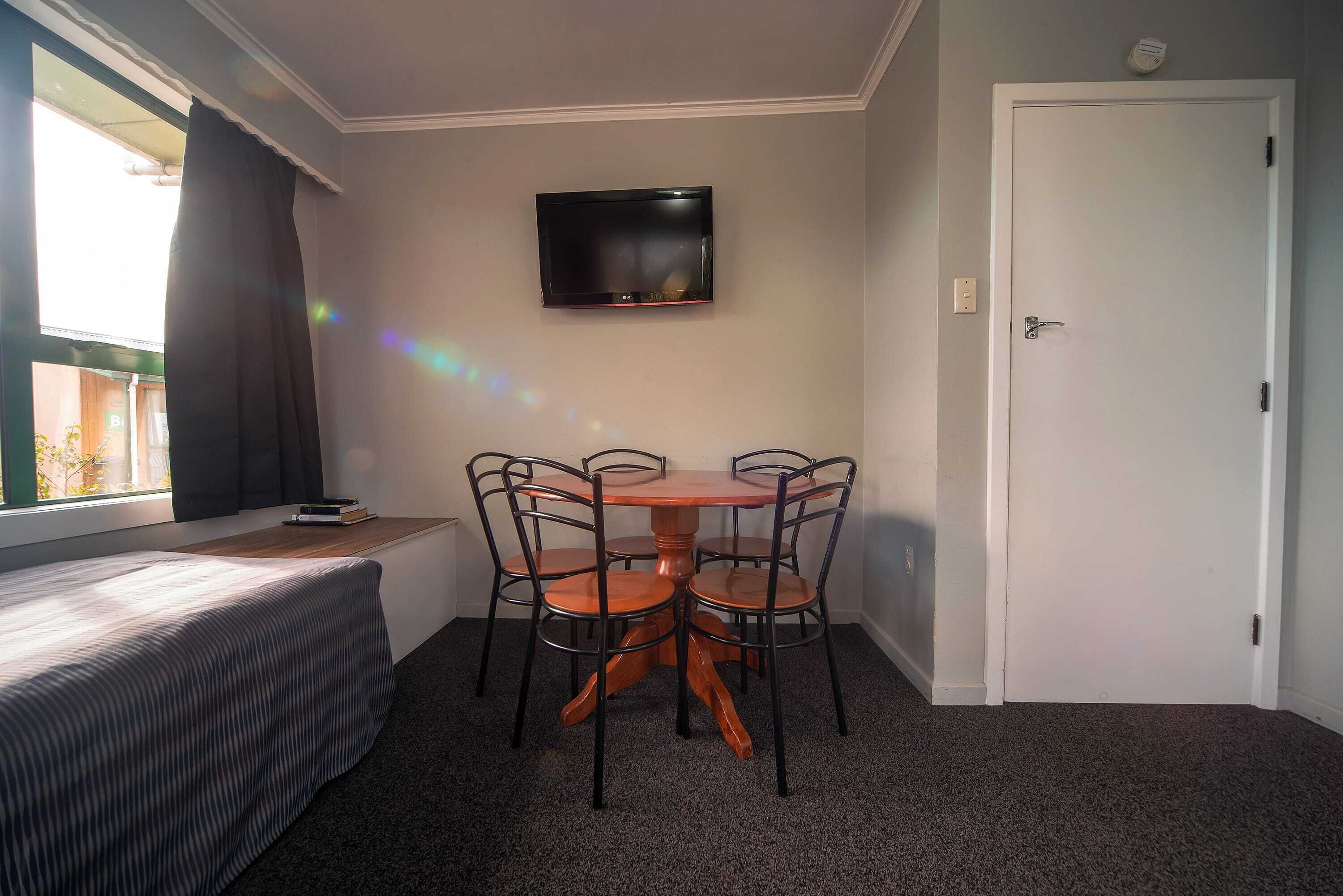 A small room with a bed, a table with four chairs, a television mounted on the wall, and a door.