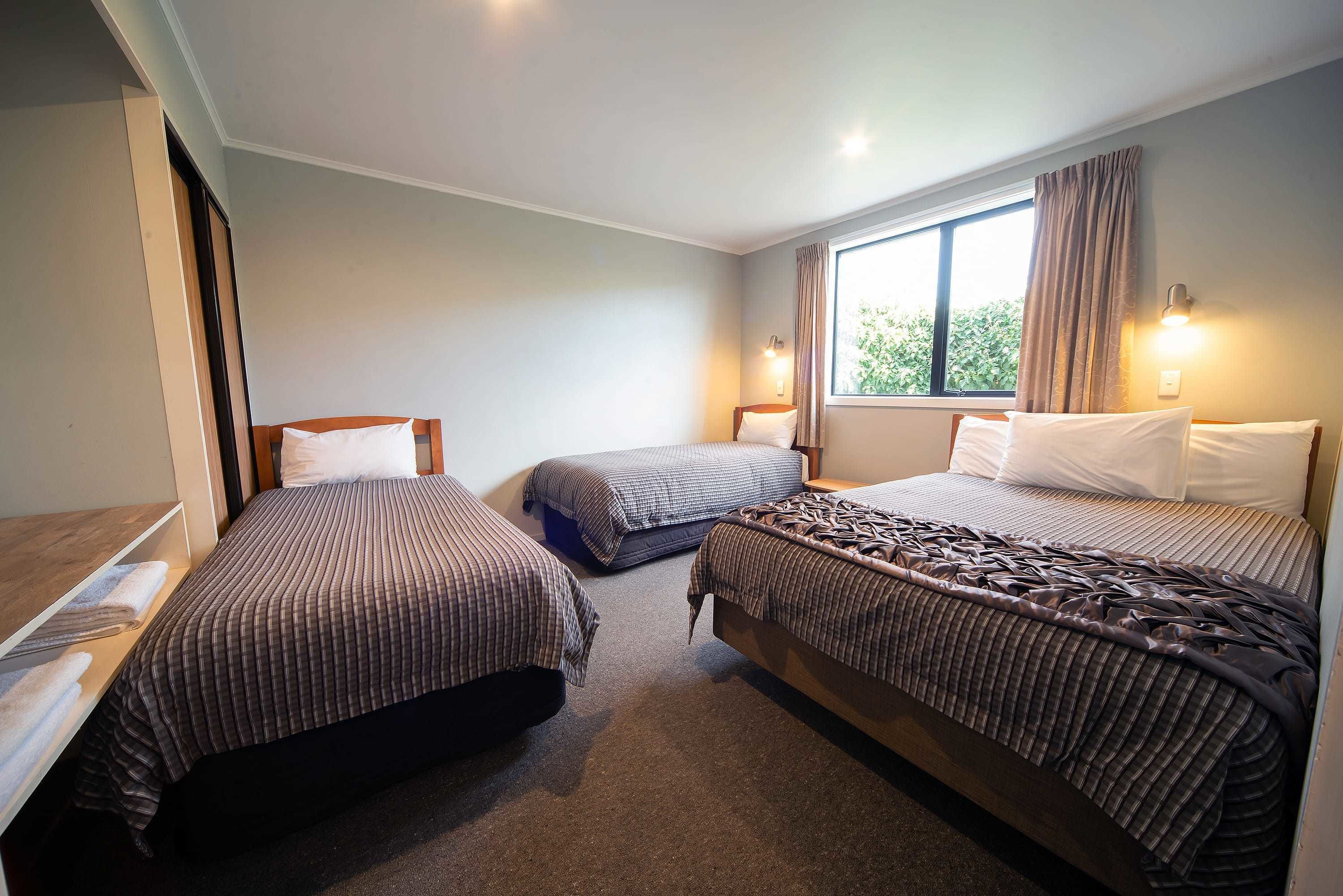 A hotel room with three beds, a window with curtains, and a bedside lamp.