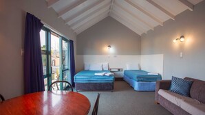 A room with a high ceiling, a bed with a blue and white striped bedspread, a sofa with a patterned cushion, a wooden table, and a chair. - Coronation Park (Ashburton)