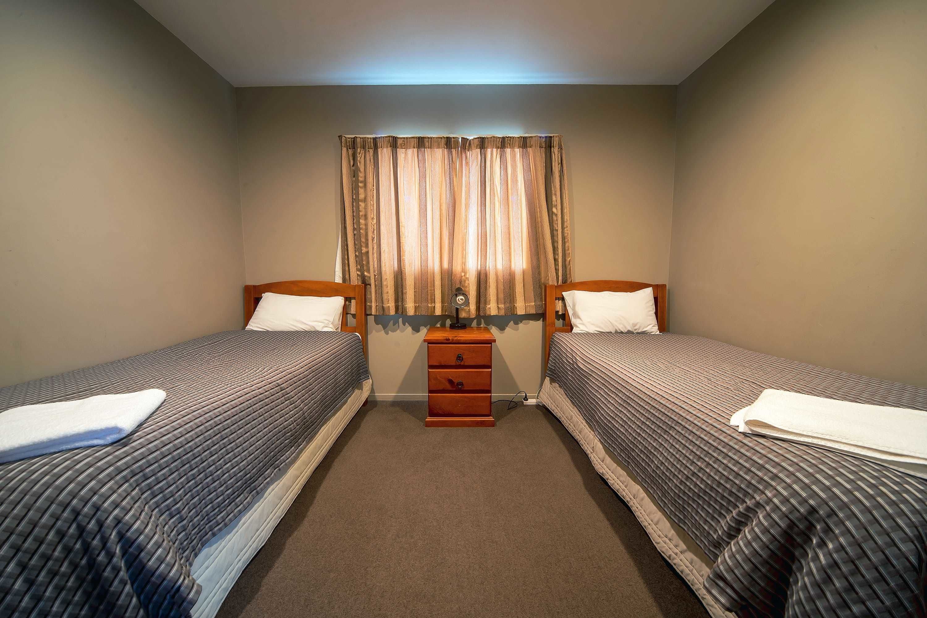 Une chambre double avec deux lits simples, une table de chevet en bois et une fenêtre avec des rideaux.