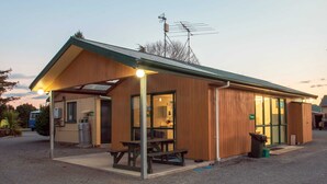 A wooden building with a covered patio, a picnic table, and a trash bin. - Coronation Park (Ashburton)