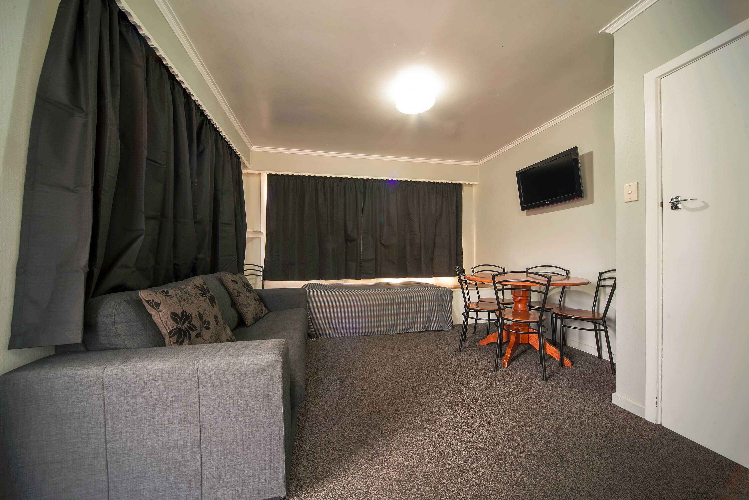 A small, furnished room with a sofa, a bed, a table with chairs, a television on the wall, and a door.