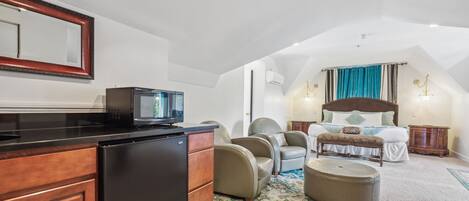 A compact, well-lit room with a kitchenette, a bed with a headboard, a seating area with chairs, and a large mirror on the wall.