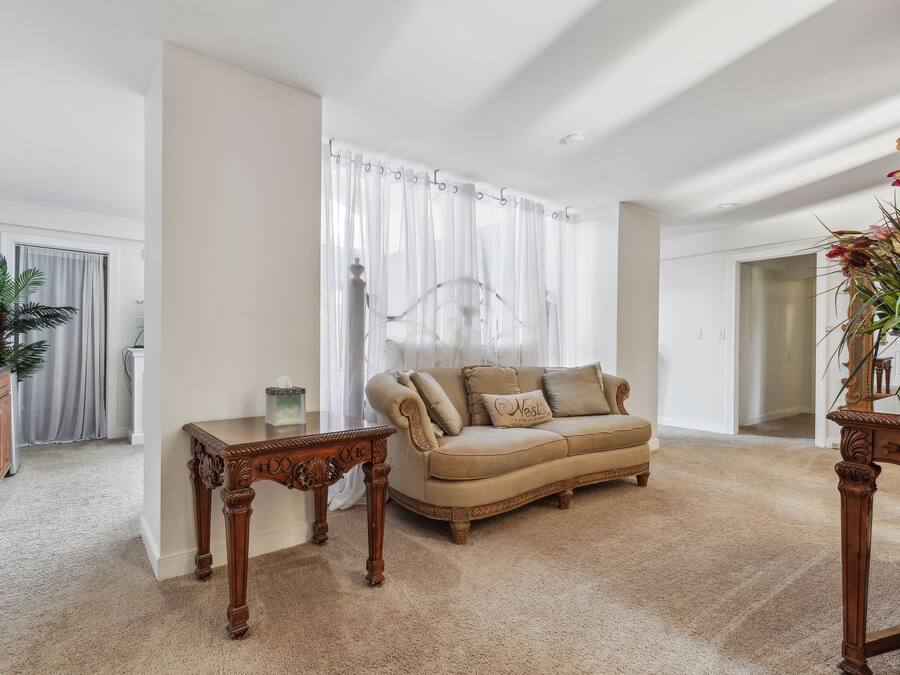 A spacious living room with a beige sofa, two wooden side tables, and a large window with sheer curtains.