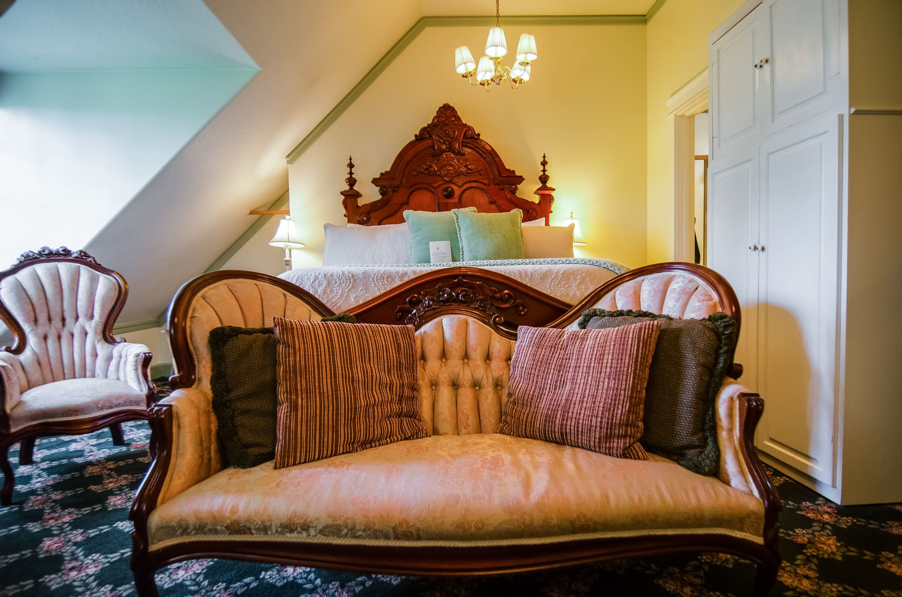 A bedroom with a large bed, a wooden headboard, a sitting area with a sofa, and a chandelier.