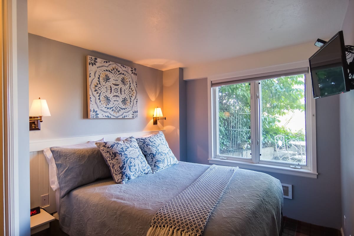 Un dormitorio con cama, dos lámparas de noche, ventana con vista a zonas verdes y televisor instalado en la pared.