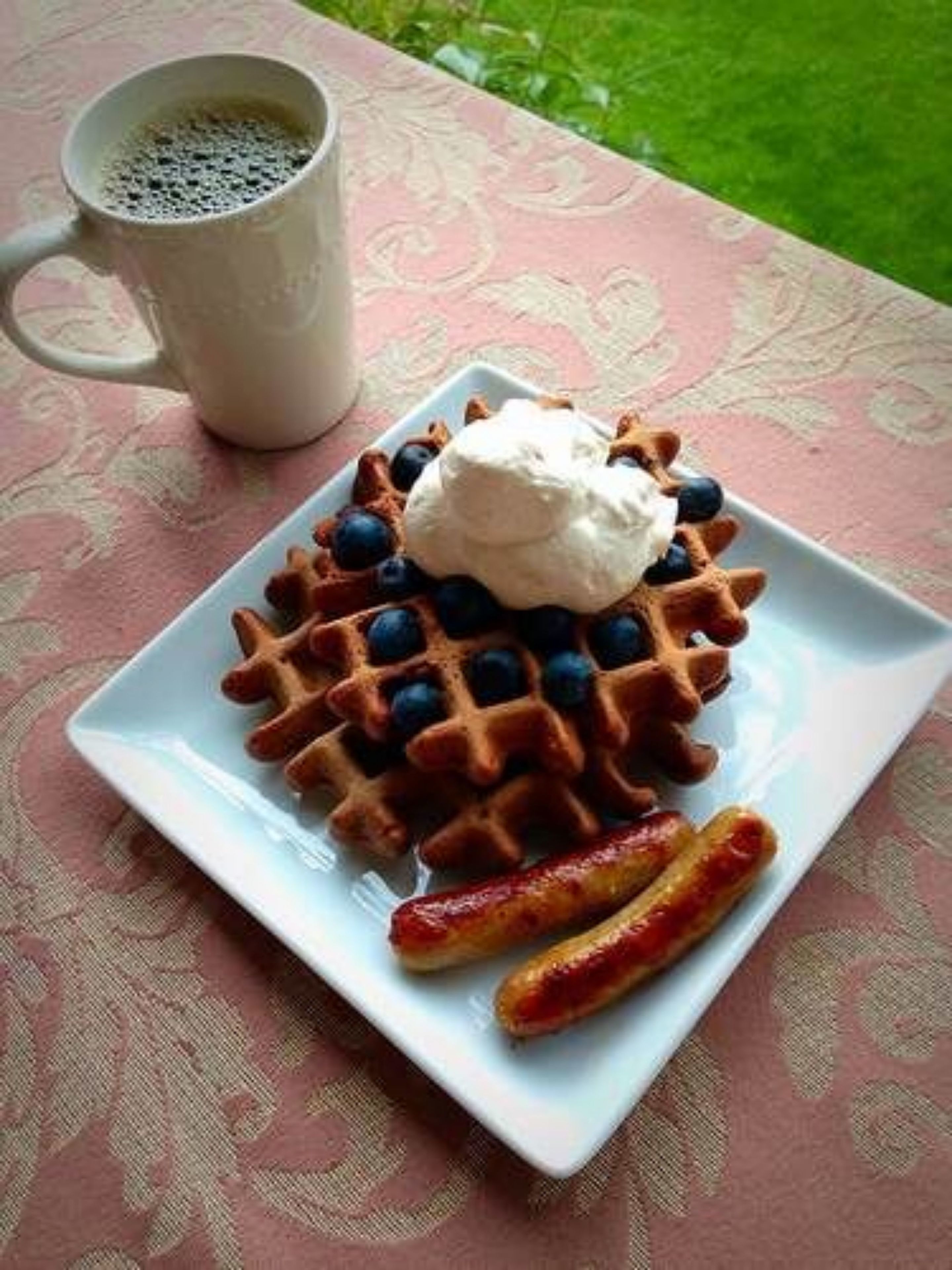 A plate with waffles topped with blueberries and whipped cream, accompanied by a mug of coffee and two sausages on a patterned tablecloth.