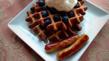 A plate with waffles topped with blueberries and whipped cream, accompanied by a mug of coffee and two sausages on a patterned tablecloth.