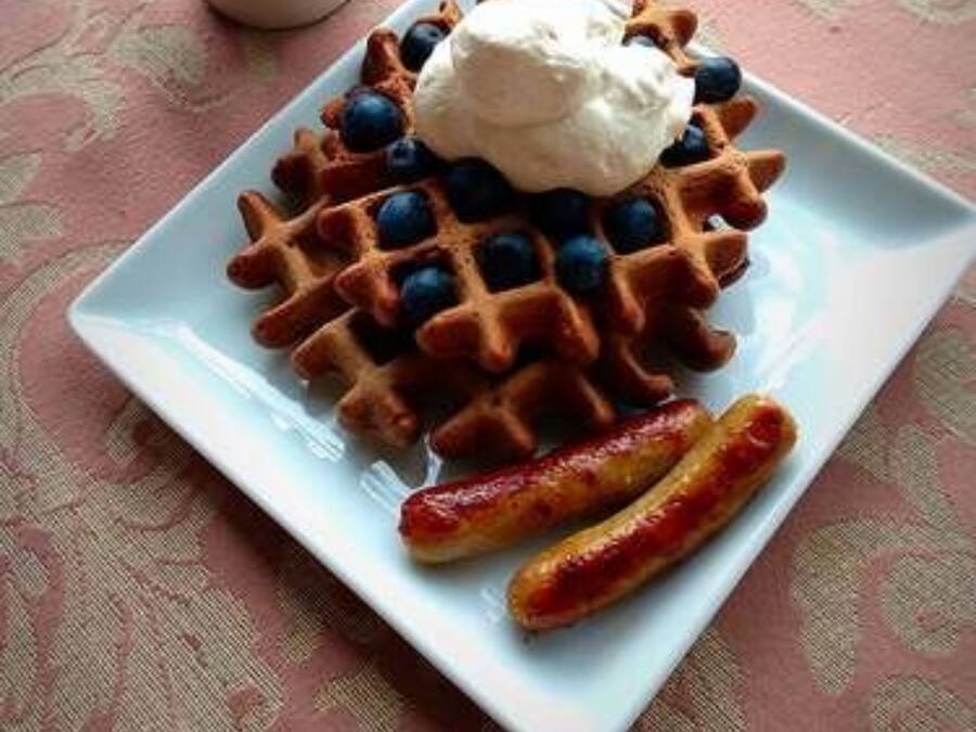 A plate with waffles topped with blueberries and whipped cream, accompanied by a mug of coffee and two sausages on a patterned tablecloth.