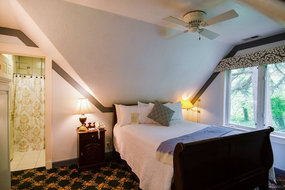 Un dormitorio con cama, mesita de noche, lámpara, ventilador de techo y ventana con vista a árboles.