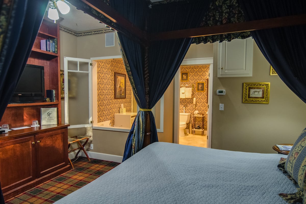 Un dormitorio con cama a dosel, cómoda de madera, televisor y baño visible a través de una puerta abierta.