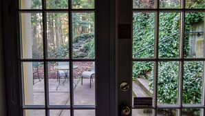 Vista a través de una ventana con celosía metálica que muestra un patio exterior con una mesa y sillas, rodeado de follaje y árboles.