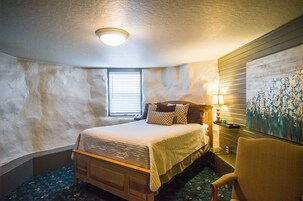 Un dormitorio con una cama de madera, una mesita de noche con una lámpara, una silla y un cuadro en la pared.