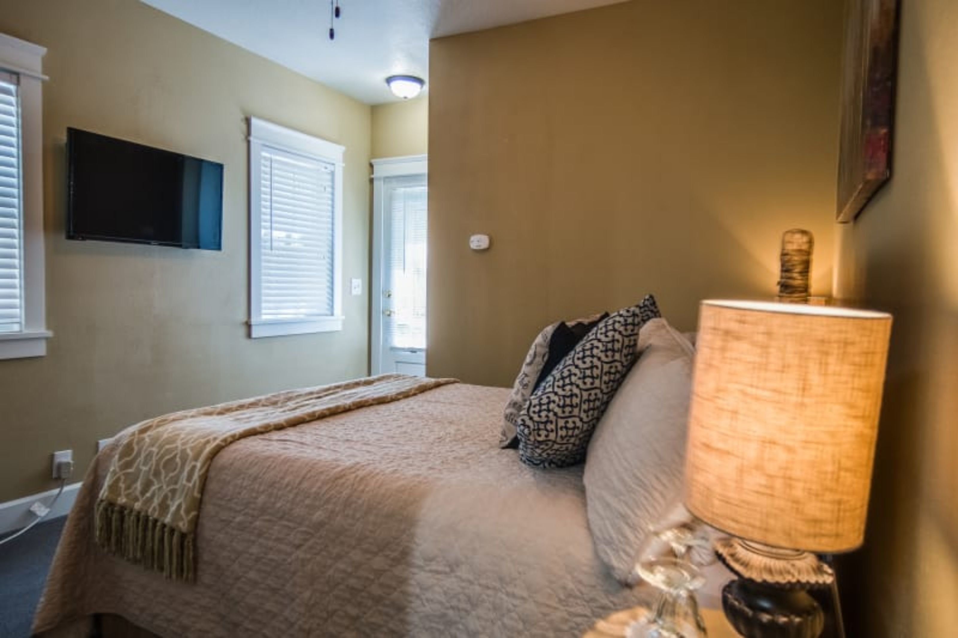 A bedroom with a bed, a lamp, a TV, and windows with blinds.