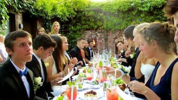 Un grup de persones assegudes al voltant d'una taula plena d'aliments i begudes, a l'aire lliure amb vegetació de fons.
