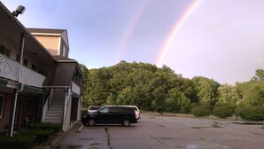 A parking lot with a building on the left and a forested hill in the background. - Budget Inn North Stonington (North Stonington)