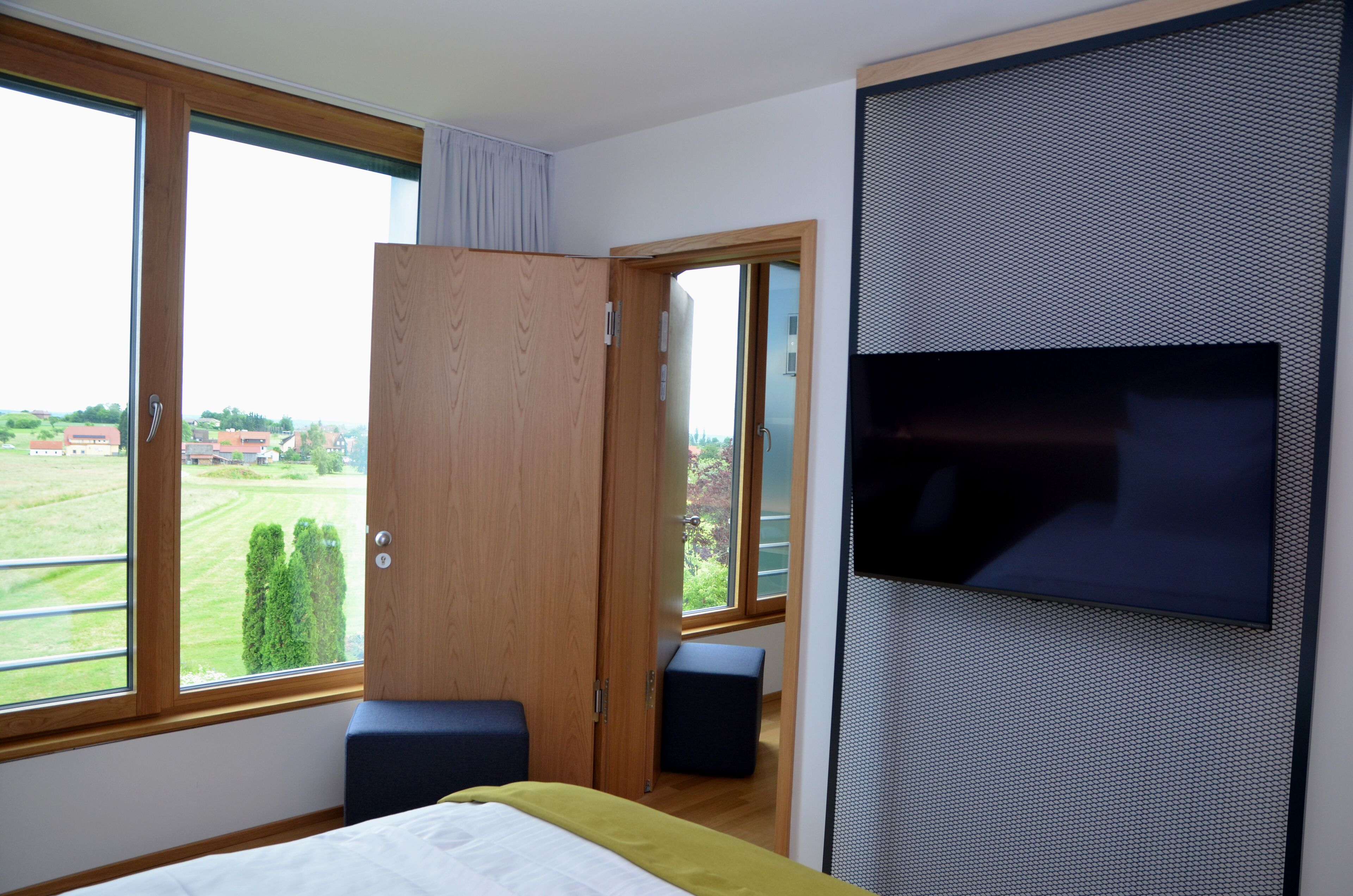 A bedroom with a large window, a bed, a TV mounted on the wall, and a wooden wardrobe.