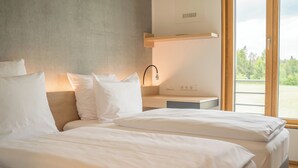 A modern hotel room with two beds, a wooden headboard, a bedside table with a lamp, and a view of greenery through large windows. - DEKRA Congresshotel Wart (Altensteig)