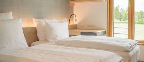 A modern hotel room with two beds, a wooden headboard, a bedside table with a lamp, and a view of greenery through large windows.