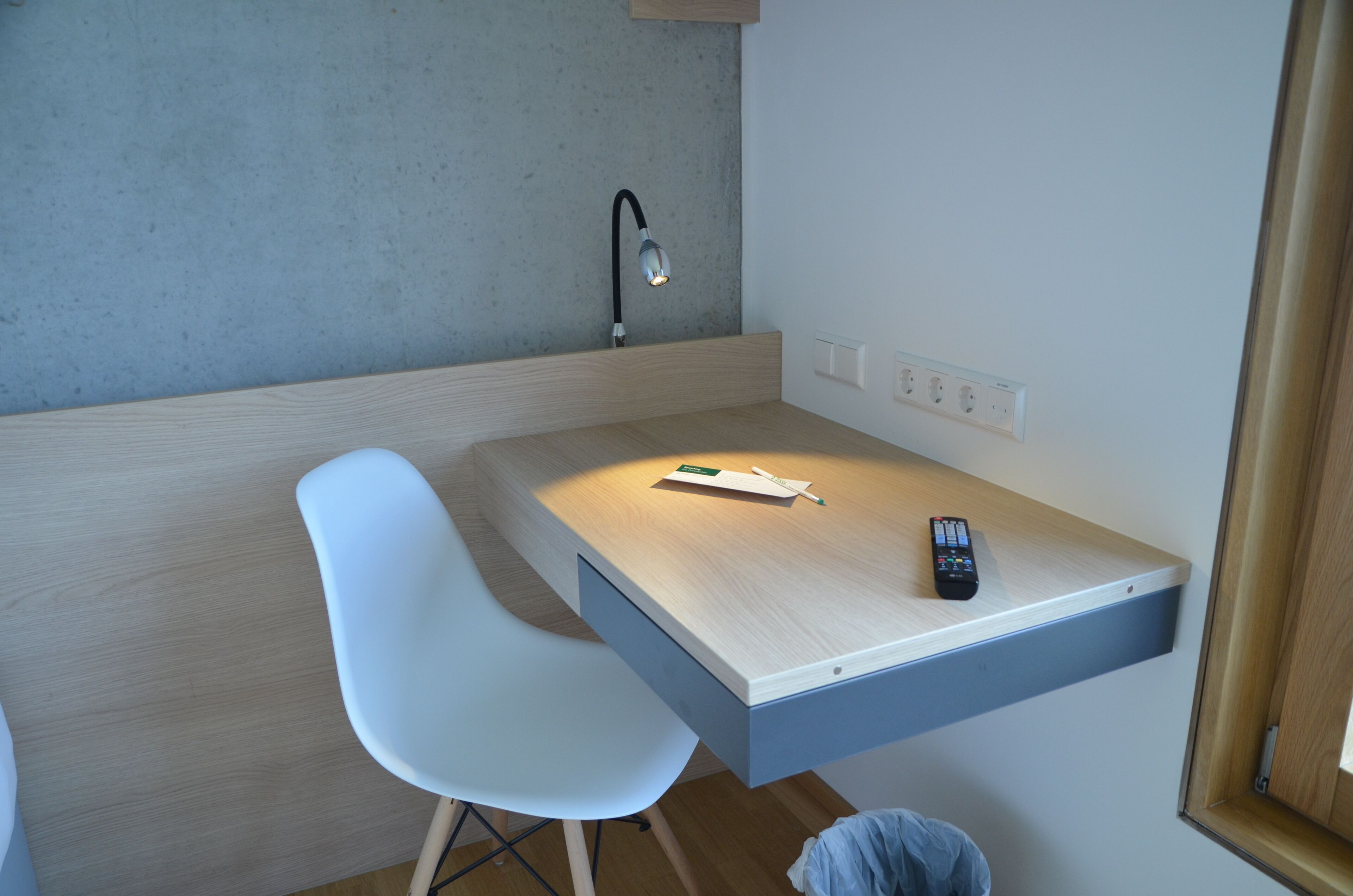 A small, modern room with a desk, a chair, a lamp, and a remote control.