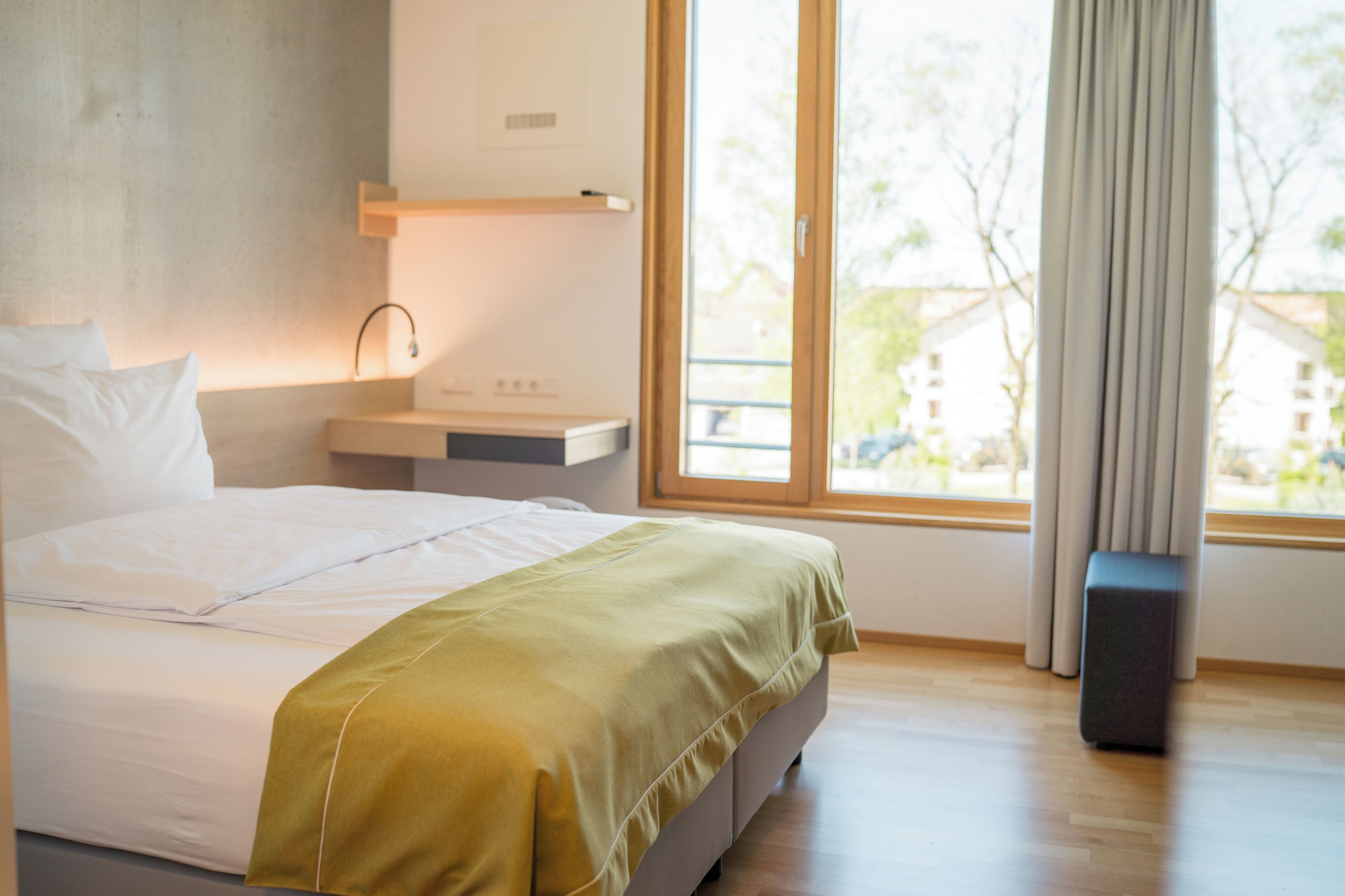 A modern hotel room with a large bed, a bedside table, a lamp, and a view of the outdoors through a large window.