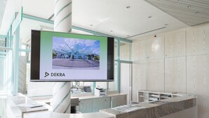 A reception desk with a large screen displaying a building image, a glass partition, and a wooden panel wall. - DEKRA Congresshotel Wart (Altensteig)