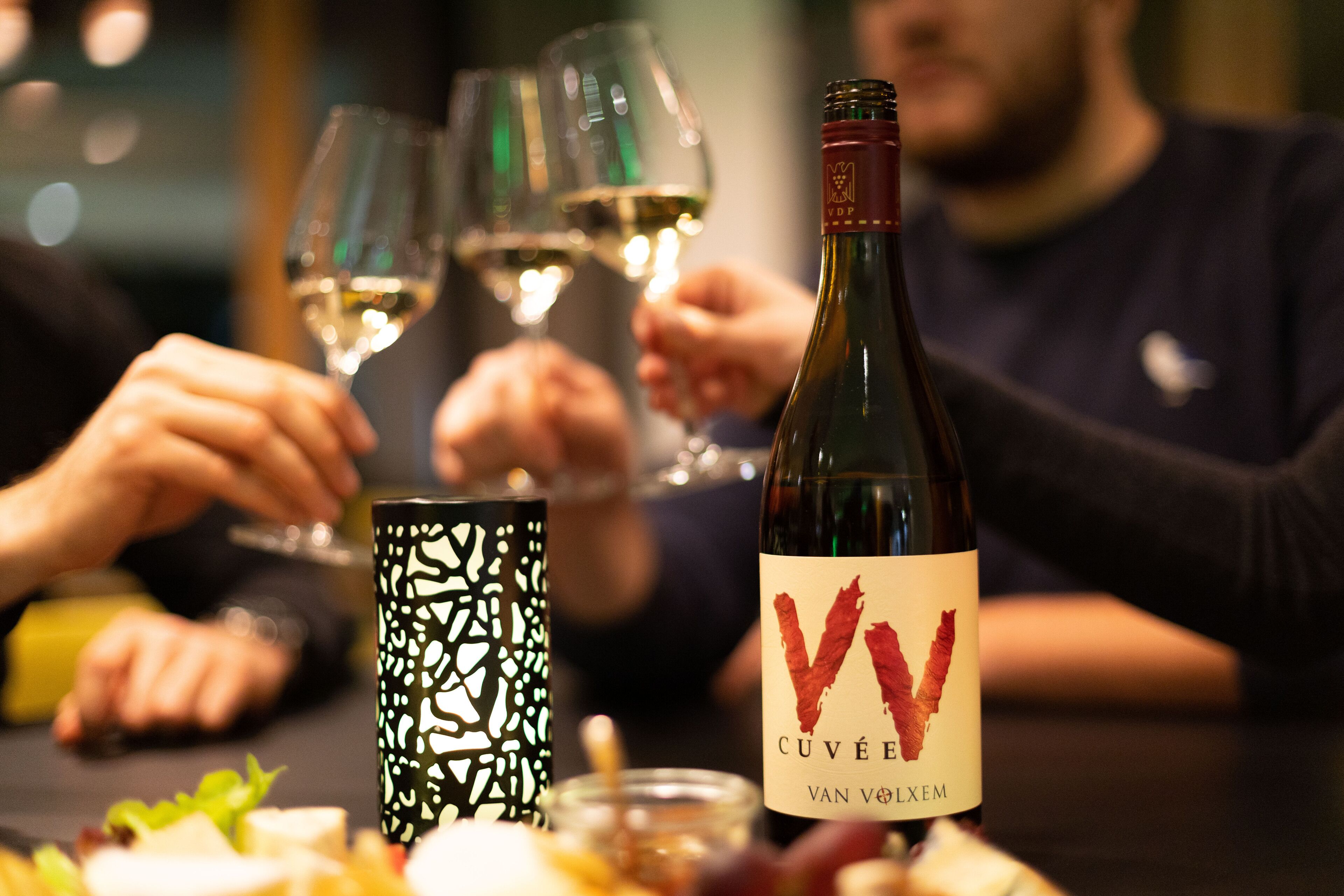 A bottle of wine with a label, glasses, and a patterned candle holder on a table.