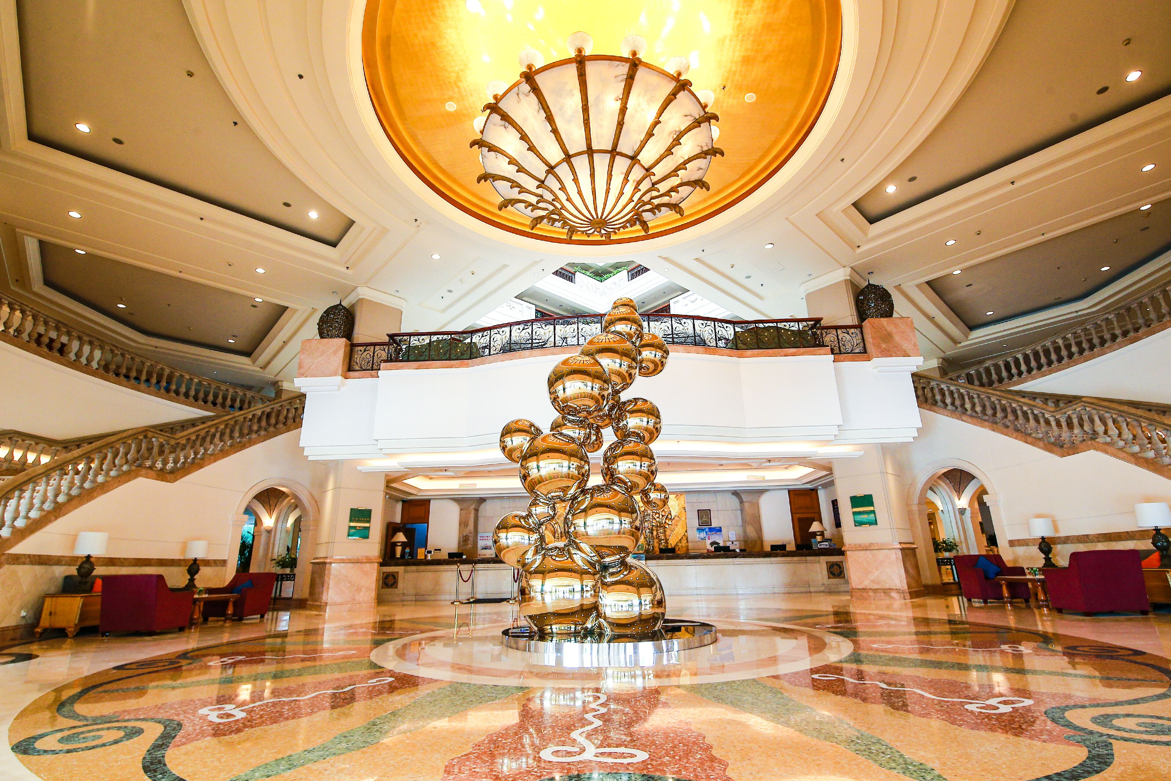 A spacious lobby with a central sculpture, a grand staircase, and a high ceiling with a decorative light fixture.