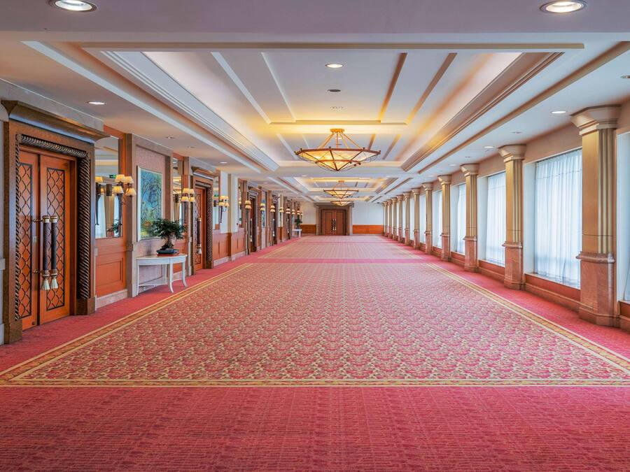 A long hallway with a red carpet, large windows, and a chandelier.