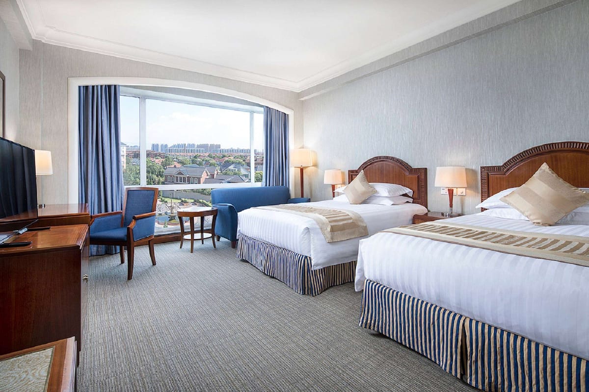 A hotel room with two beds, a desk, a chair, and a large window offering a city view.