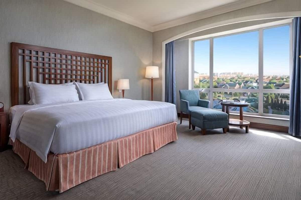 A hotel room with a large bed, a small table, a chair, and a view of a cityscape through large windows.