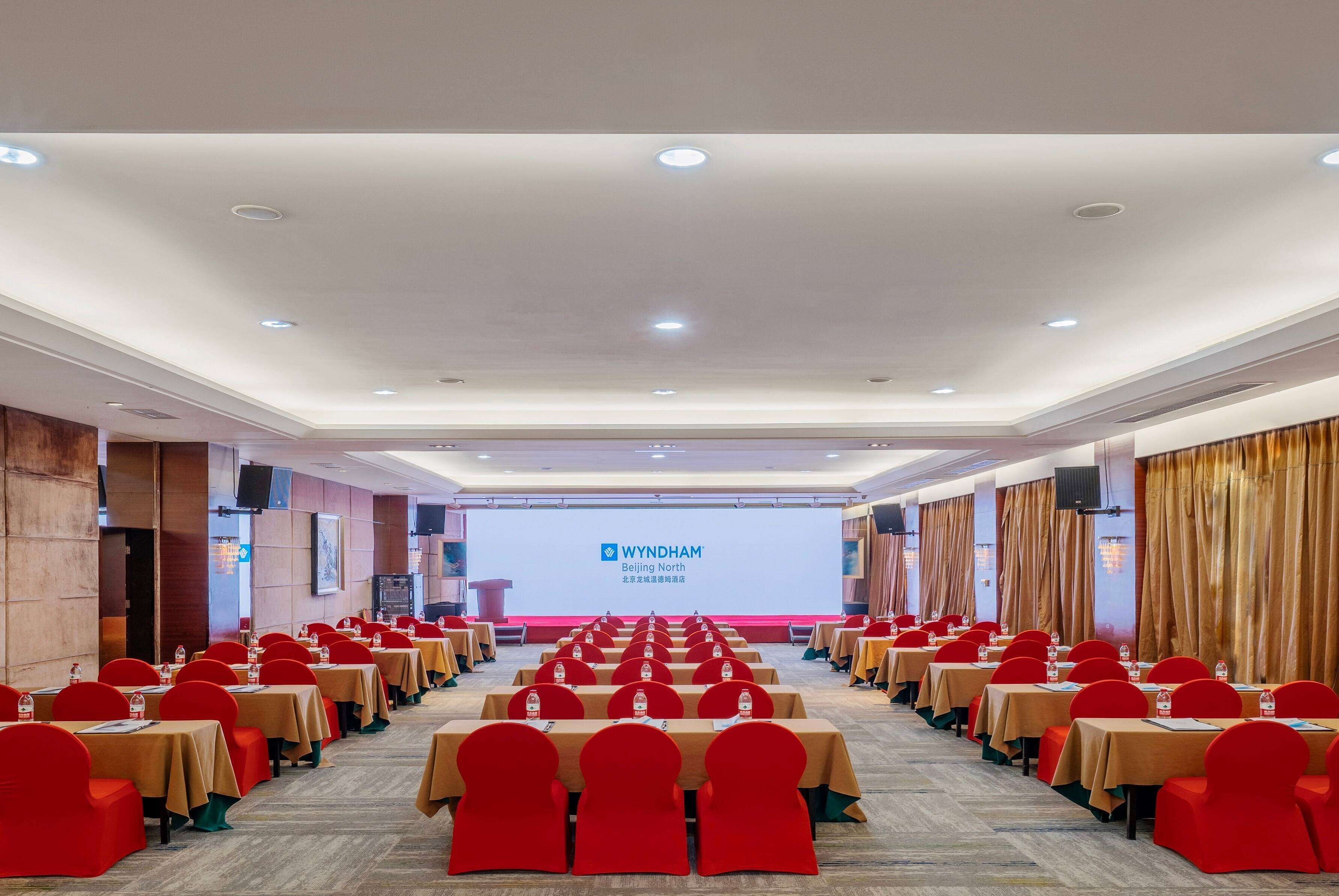 Ett konferensrum med rader av bord och stolar, en stor skärm som visar bilder från WYNDHAM Beijing North och en scen med ett talarstånd.