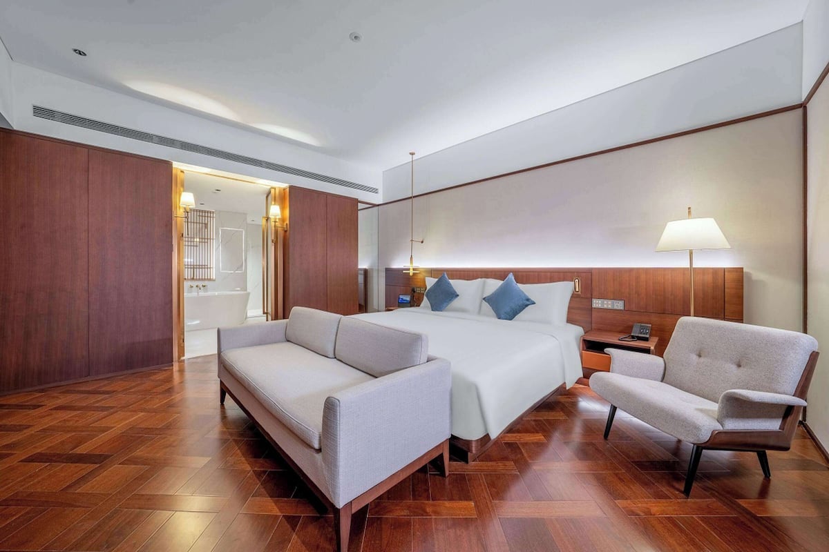 A modern hotel room with a large bed, a sofa, a chair, a nightstand, and a lamp.