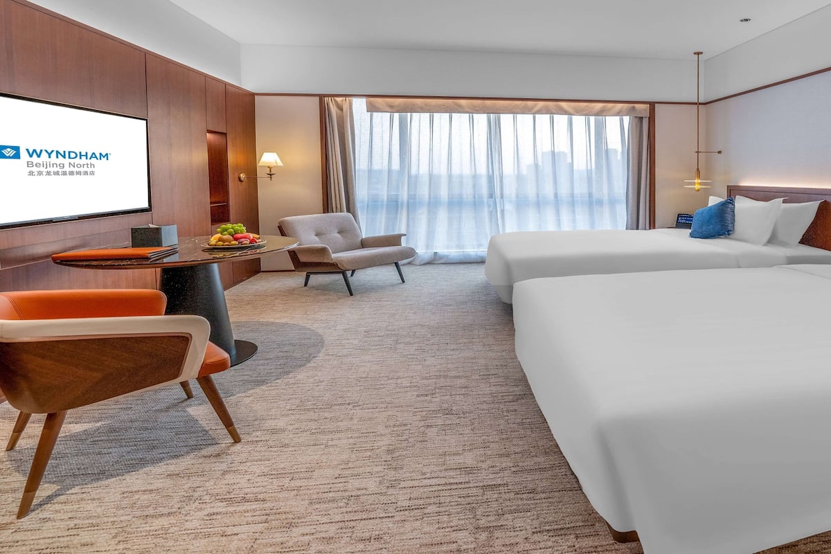 A hotel room with a bed, a desk with a laptop, a chair, a TV screen, and a large window with curtains.