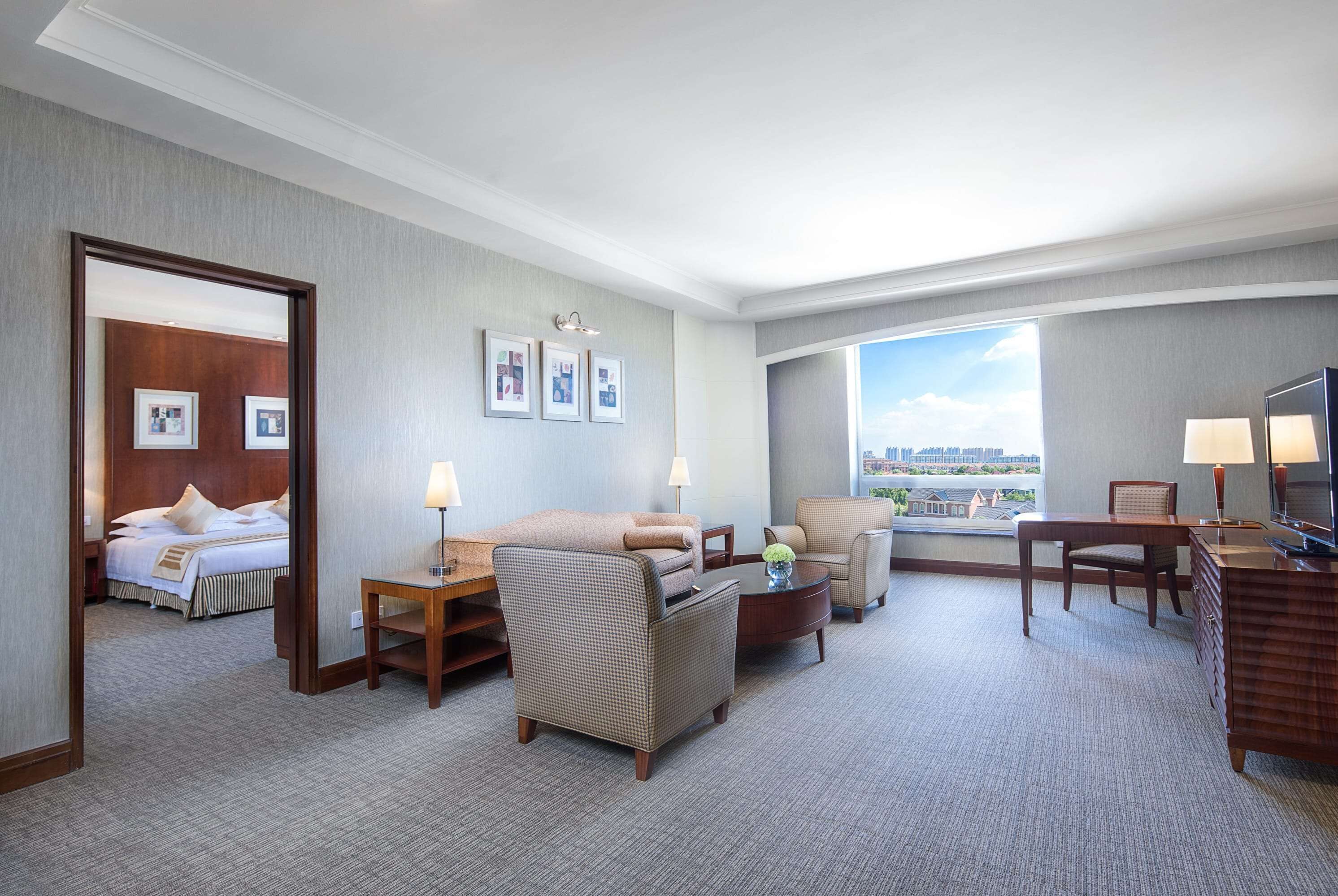 Une chambre d’hôtel avec un lit, un canapé, un bureau et une chaise. Il y a trois tableaux encadrés au mur et une fenêtre donnant sur des immeubles.