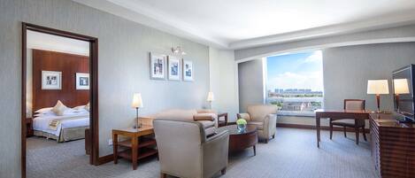 Une chambre d’hôtel avec un lit, un canapé, un bureau et une chaise. Il y a trois tableaux encadrés au mur et une fenêtre donnant sur des immeubles.