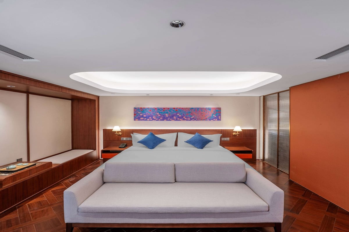 A modern hotel room with a large bed, a sofa, a wooden bench, and a colorful abstract painting on the wall.