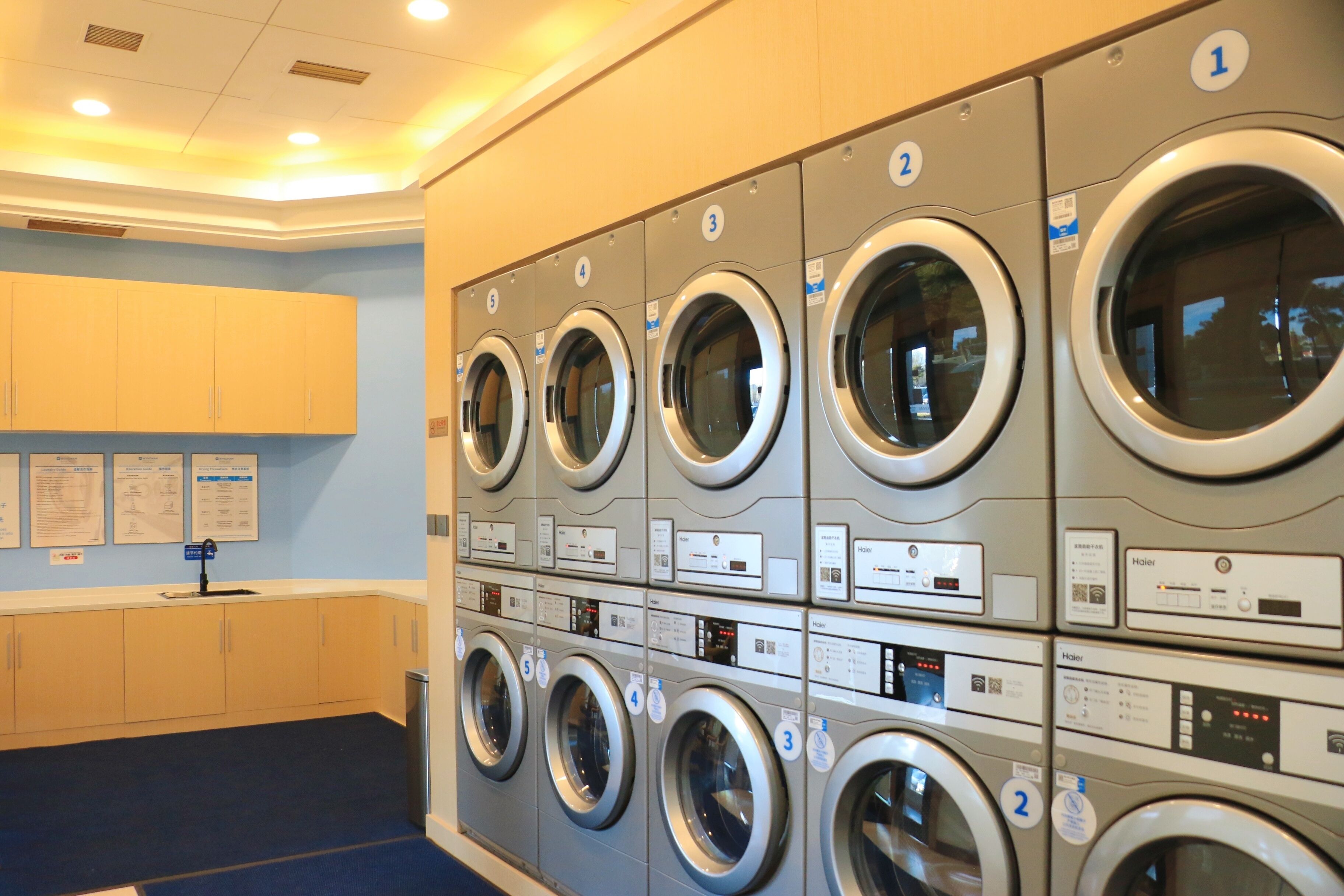 A row of six stacked front-loading washing machines with control panels, a sink, and informational posters on the wall.