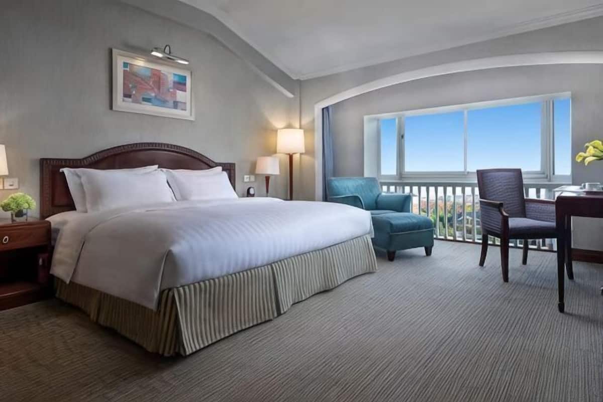A hotel room with a large bed, a desk with a chair, a balcony with a view, and a window.