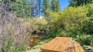 Una mesa de picnic de madera en un entorno natural, con un arroyo y follaje denso al fondo.