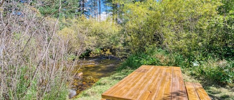 Una mesa de picnic de madera en un entorno natural, con un arroyo y follaje denso al fondo.