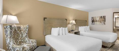 A hotel room with two beds, a chair, a desk, and a lamp.