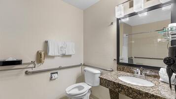 Baño con inodoro, lavamanos y secador de manos. Cuenta con barras de apoyo y un perchero para toallas.