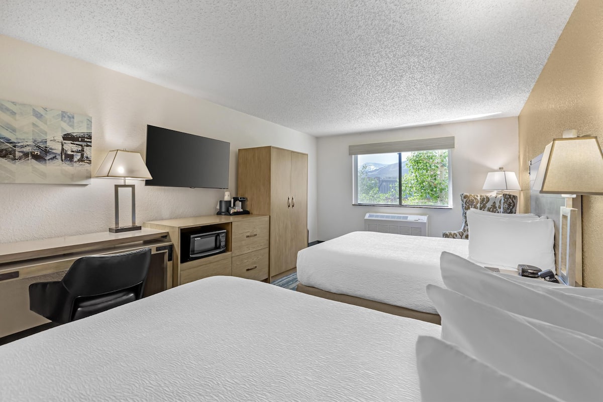 A hotel room with two beds, a desk with a television, a microwave, and a window with a view of greenery.