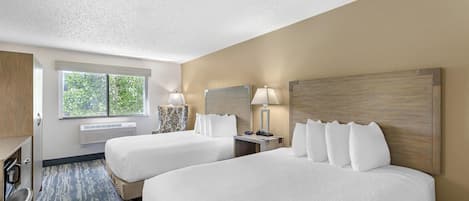 A hotel room with two beds, a desk, a chair, a window, and a wall-mounted heater.