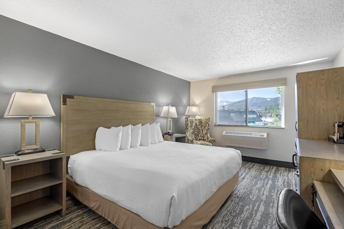A hotel room with a large bed, two bedside tables with lamps, a chair, a desk, and a window with a view of mountains.