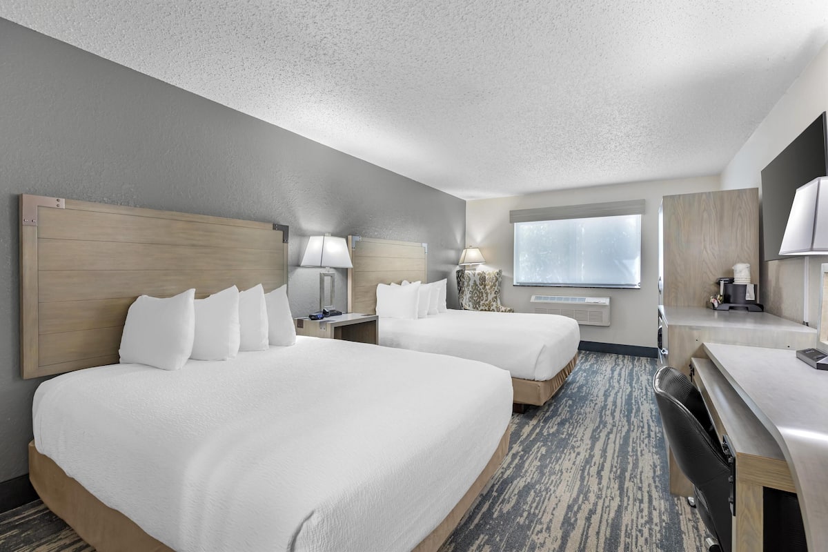 A hotel room with two beds, a desk, a chair, a window, and a lamp.