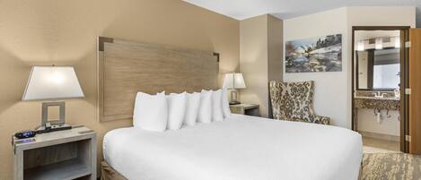 A hotel room with a large bed, two bedside tables with lamps, a chair, a painting, and a bathroom mirror.