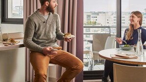 A modern hotel room with a large window offering a city view, a dining area with a round table, and a kitchenette.