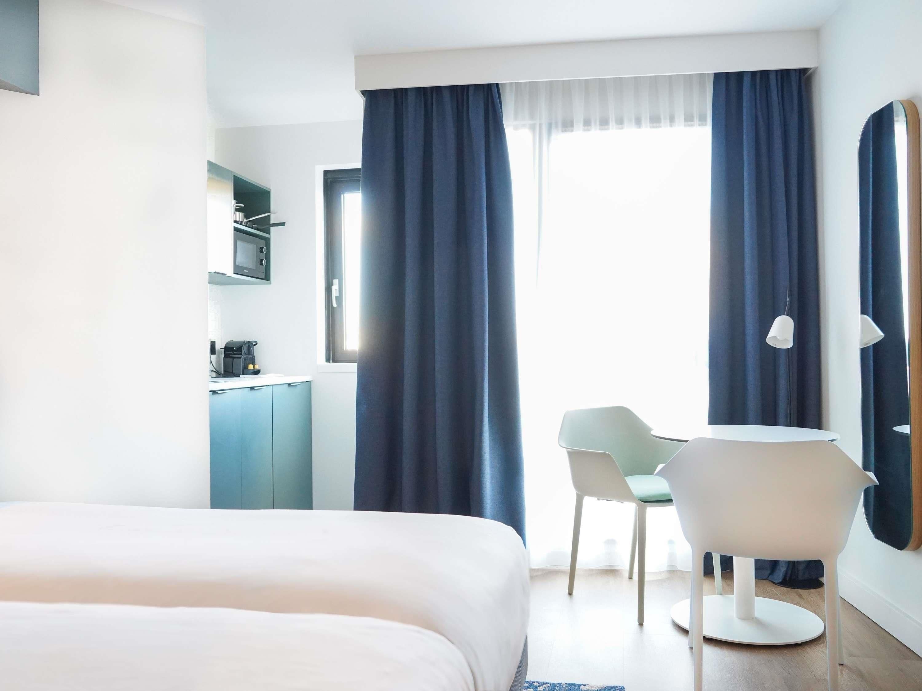 Una habitación de hotel con una cama, un escritorio con silla, un espejo y un ventanal con cortinas.